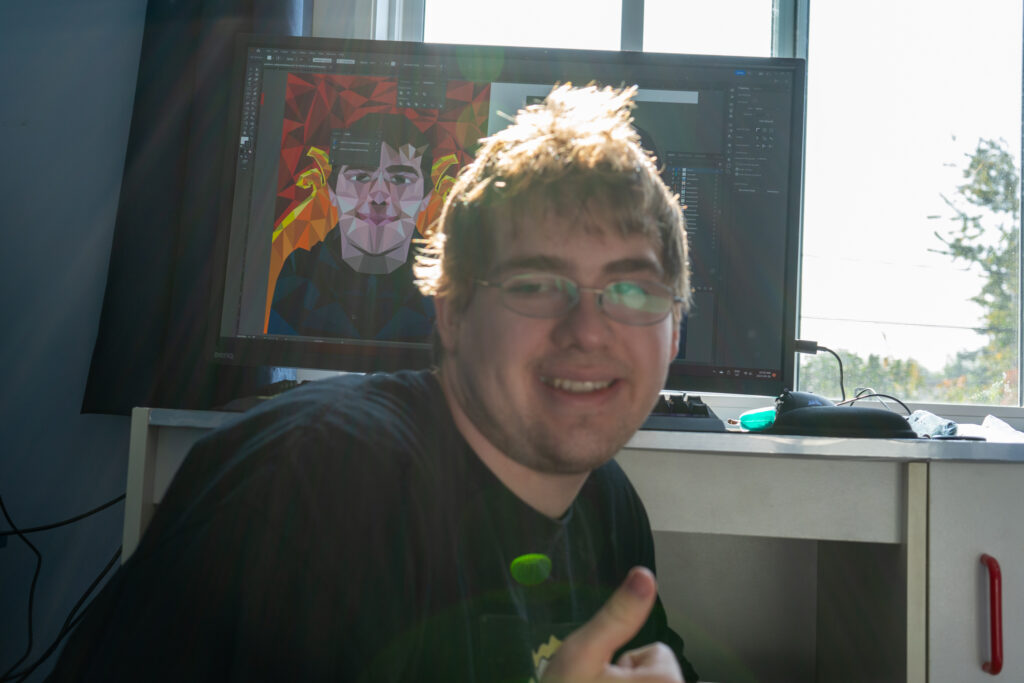 Picture of Max Parker in front of a desk and computer screen showing his work, smiling and giving a thumbs up.